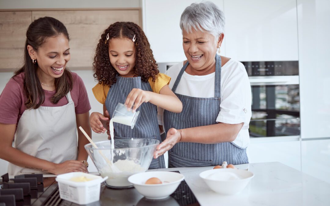 Three generations of women cooking together in the kitchen with young girl pouring liquid in bowl