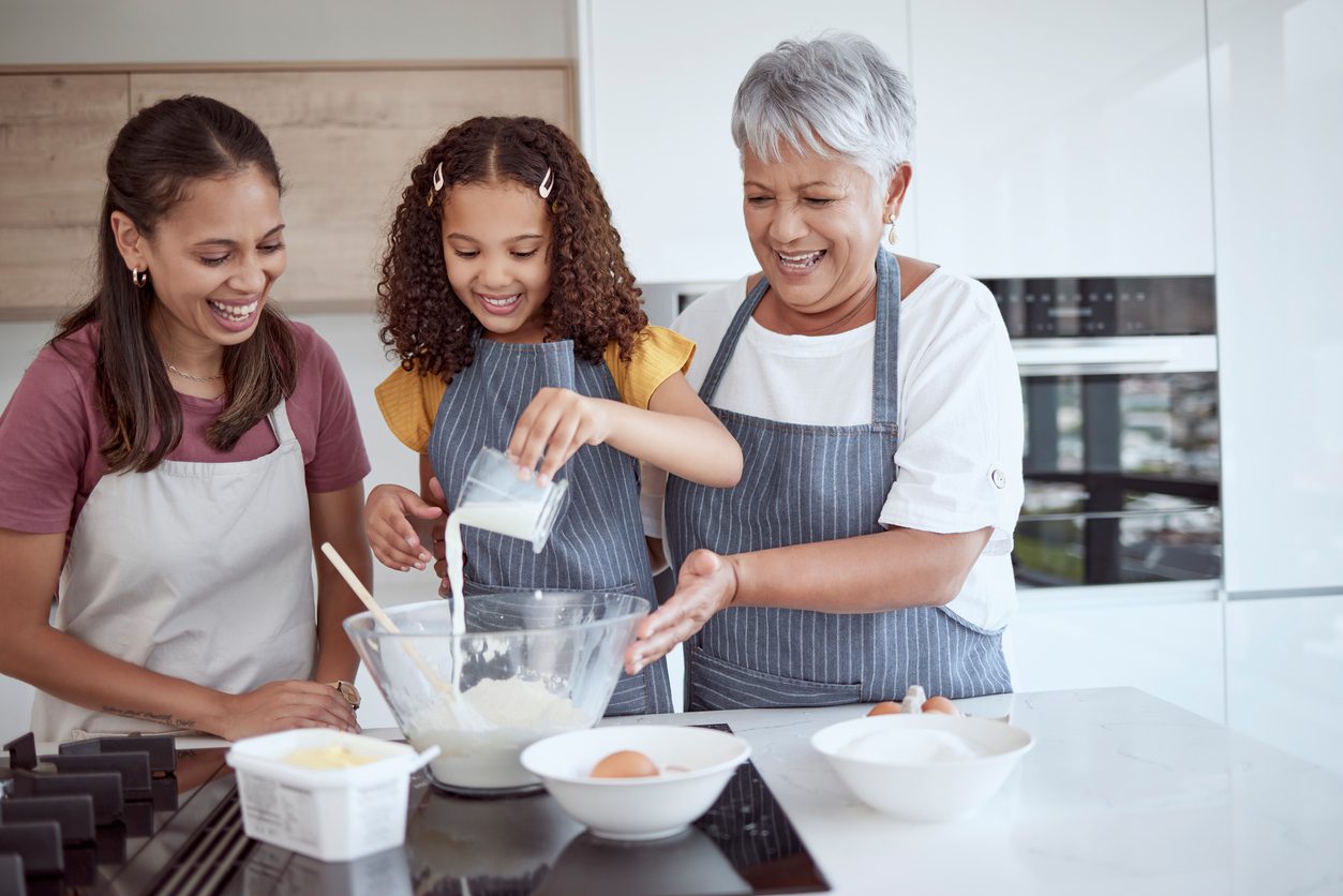 Three generations of women cooking together in the kitchen with young girl pouring liquid in bowl