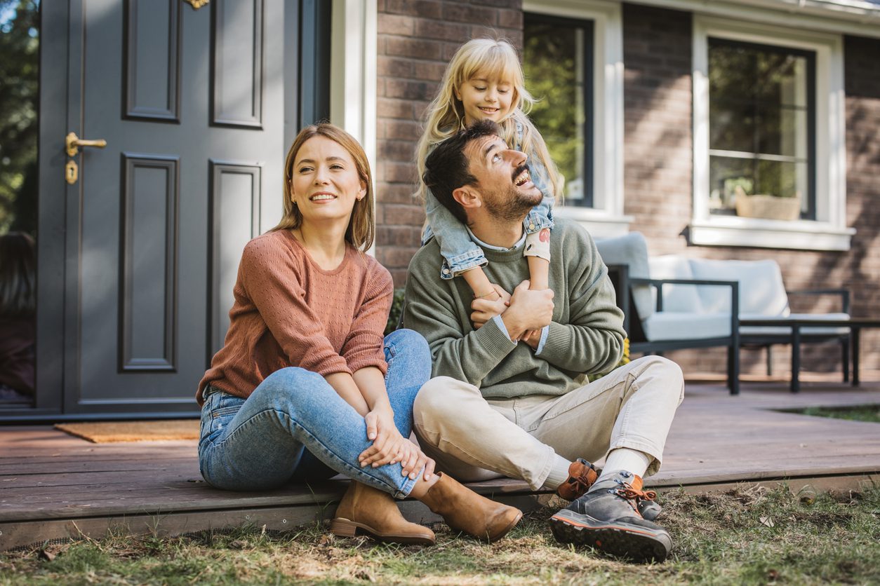 Mother and father on front porch step with young daughter hugging dad from behind